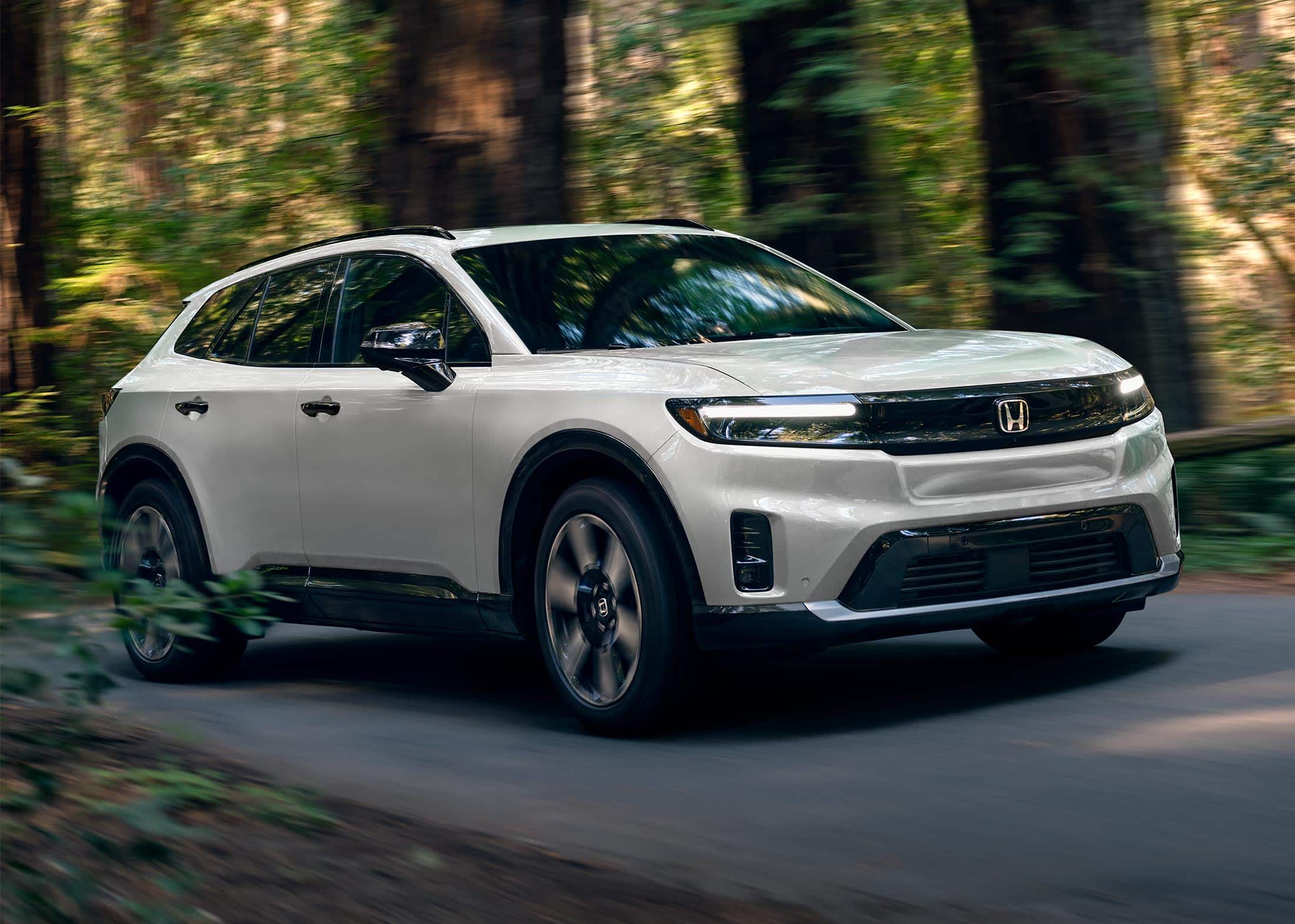 3/4 front passenger view of the 2024 Honda Prologue Elite in Snowfall Pearl, shown on a paved road with redwood trees behind it.