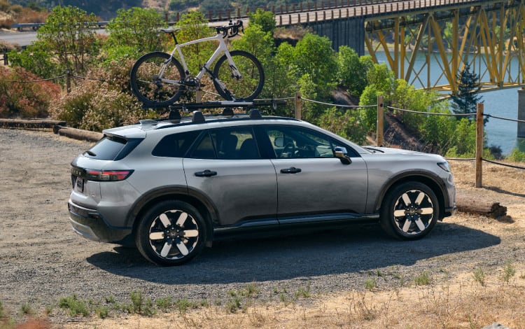 2026 Honda Prologue Elite in Frostline White Pearl, parked at a bridge overlook near a lake, with one bike attached to the roof, featuring Honda Genuine Accessories.